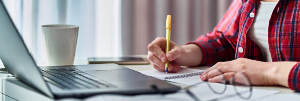 A person in a red plaid shirt writes in a notebook at a desk with a laptop, papers, and a coffee cup.