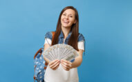 Portrait of young smiling attractive woman student in denim clothes with backpack holding lots of dollars